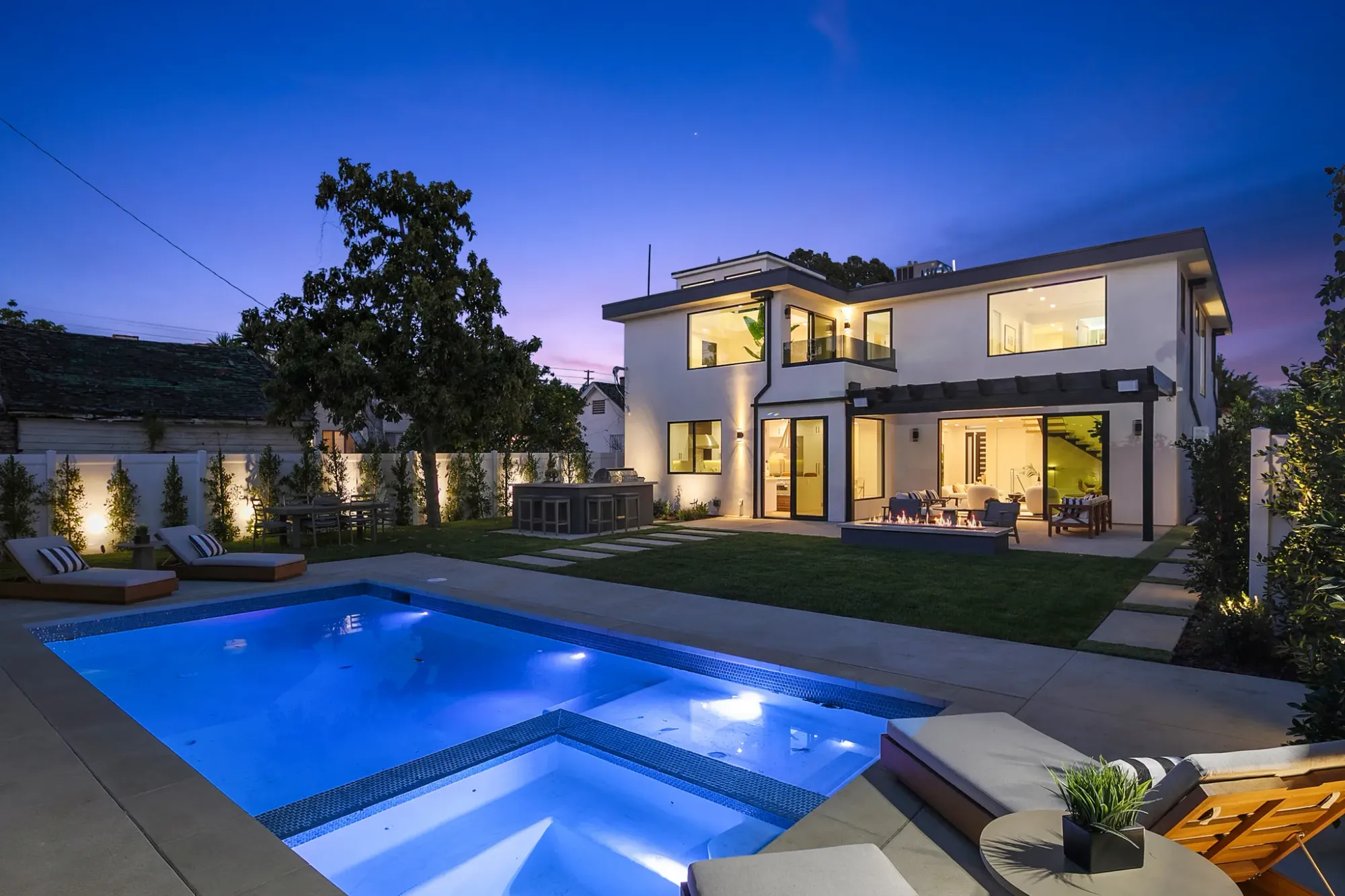 Lit pool and spa behind a new two-story white home at dusk